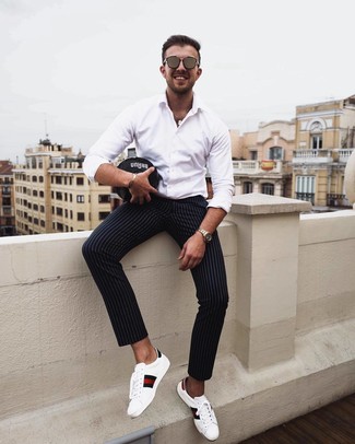 Модный лук: белая рубашка с длинным рукавом, черные брюки чинос в вертикальную полоску, белые кожаные низкие кеды с принтом, коричневые солнцезащитные очки