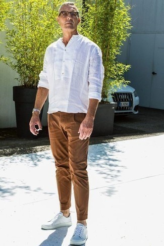 С чем носить темно-коричневые брюки чинос за 50 лет: Белая льняная рубашка с длинным рукавом и темно-коричневые брюки чинос — отличный выбор, если ты хочешь создать расслабленный, но в то же время модный мужской лук. Ты сможешь легко адаптировать такой ансамбль к повседневным условиям городской жизни, завершив его белыми кожаными низкими кедами.