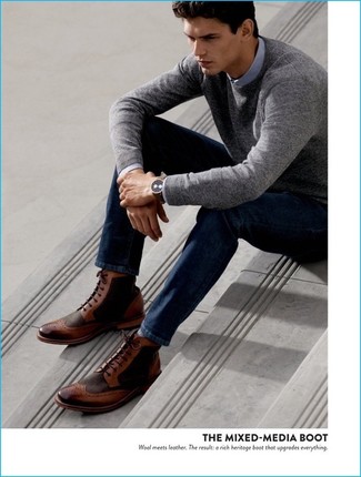 Как носить темно-синие джинсы с темно-коричневыми кожаными ботинками броги: Серый свитер с круглым вырезом и темно-синие джинсы — неотъемлемые предметы в гардеробе джентльменов с превосходным вкусом в одежде. Такой образ получает новое прочтение в тандеме с темно-коричневыми кожаными ботинками броги.