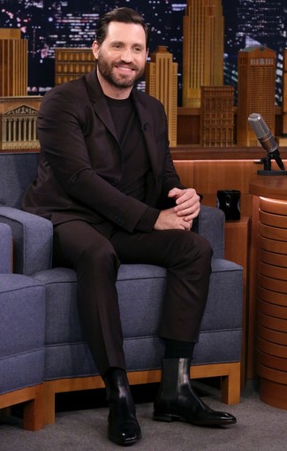Модный лук: темно-коричневый костюм, черный свитер с круглым вырезом, черные кожаные ботинки челси, черные носки