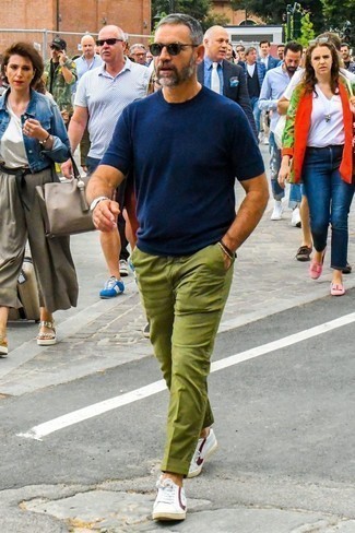 С чем носить темно-зеленые брюки чинос в жару: Темно-синяя футболка с круглым вырезом в сочетании с темно-зелеными брюками чинос не прекращает импонировать стильным молодым людям. Что же до обуви, бело-красные кожаные низкие кеды — наиболее подходящий вариант.